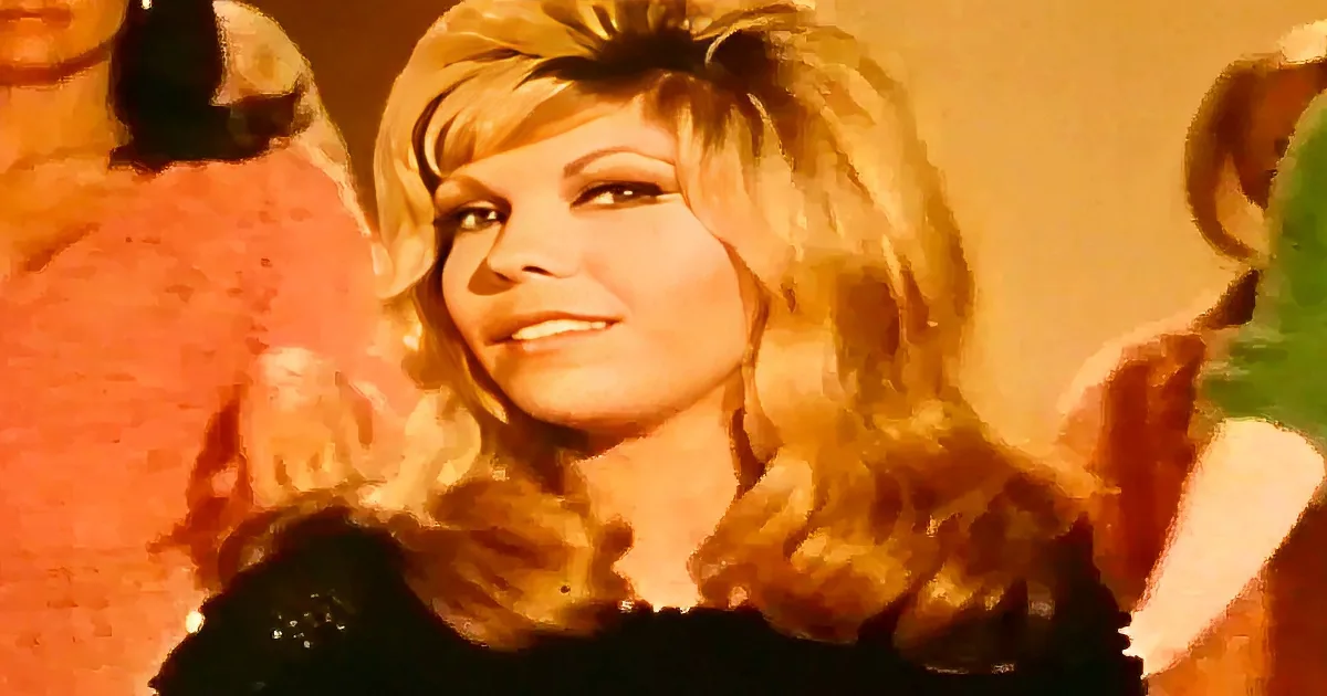 A woman with blonde, voluminous hair and a black top is smiling.