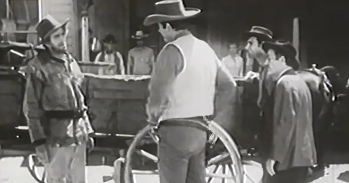 A cowboy in a white vest and hat stands near a wagon, surrounded by other men in western attire.