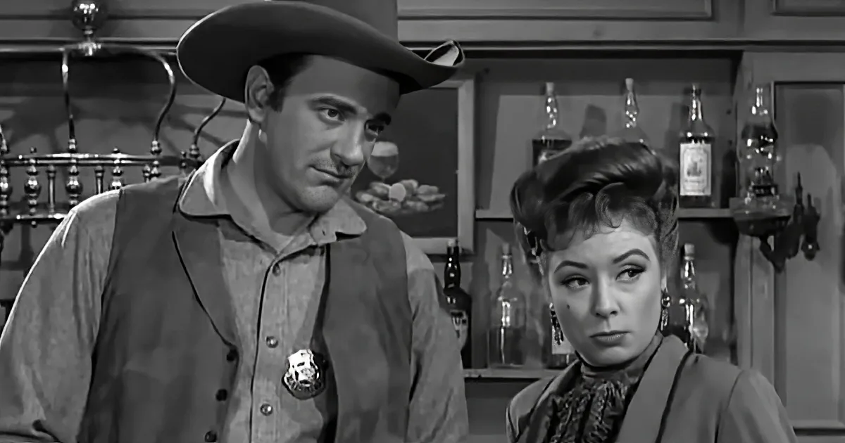A man in a cowboy hat and vest stands next to a woman in a Western setting with bottles in the background.