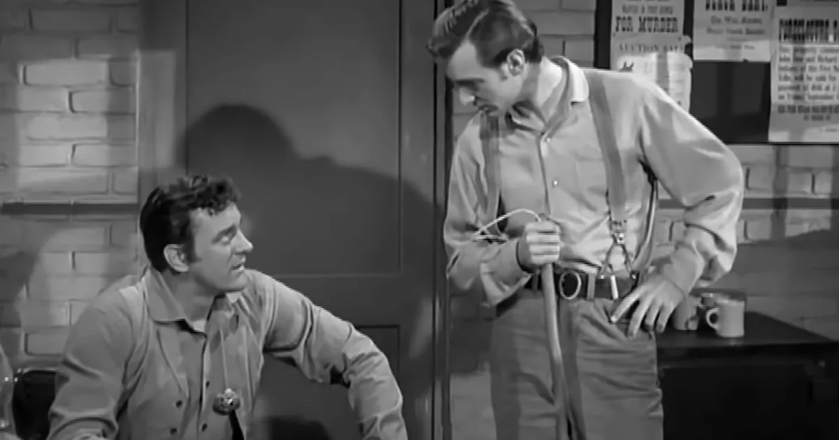 A man in a light shirt and suspenders holds a rope, while another man in a similar shirt leans forward, both in a room...