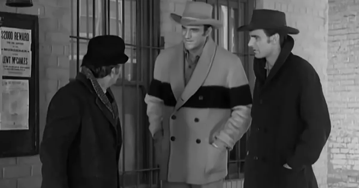 Three men in hats and coats stand in front of a jail cell, one in a striped coat appears to be speaking.