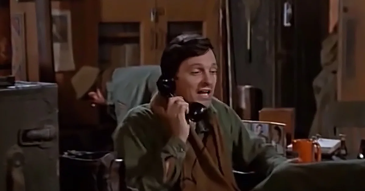 A man in a green jacket is talking on a black phone in a cluttered room.