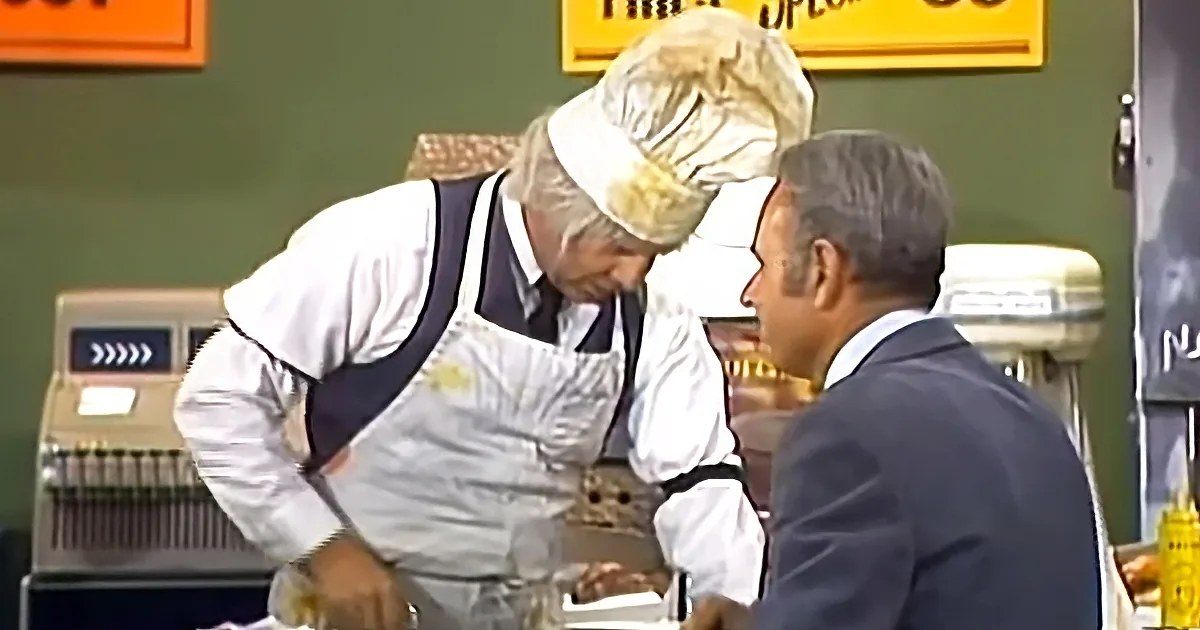 A chef in a white uniform and hat is leaning over a counter, interacting with a man in a suit.
