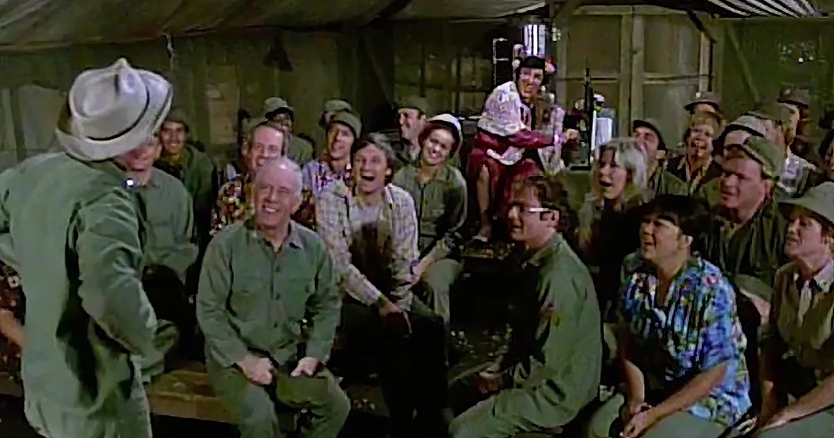 A man in a green military uniform and hat is addressing a group of soldiers and civilians in a tent.