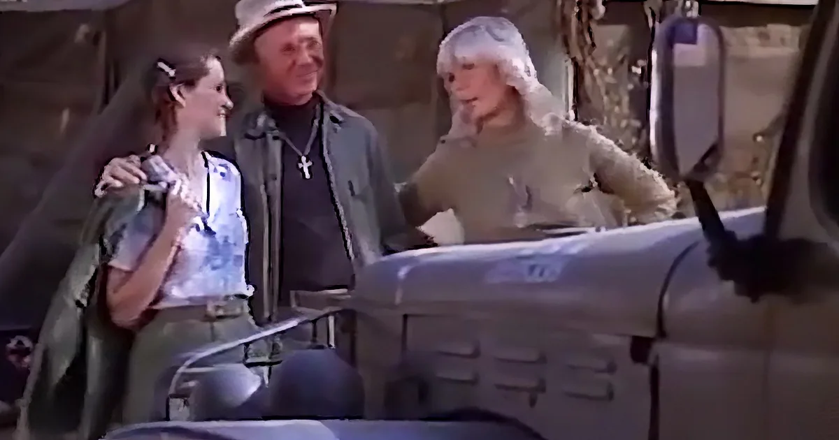 A man in a hat and jacket stands next to a woman in a white shirt and green vest, both looking at a woman driving a vehicle.
