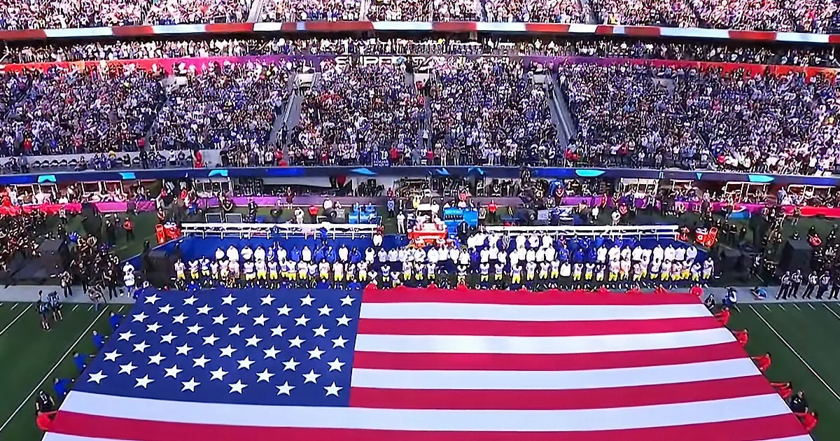 Players stand in formation behind a large American flag on a football field.