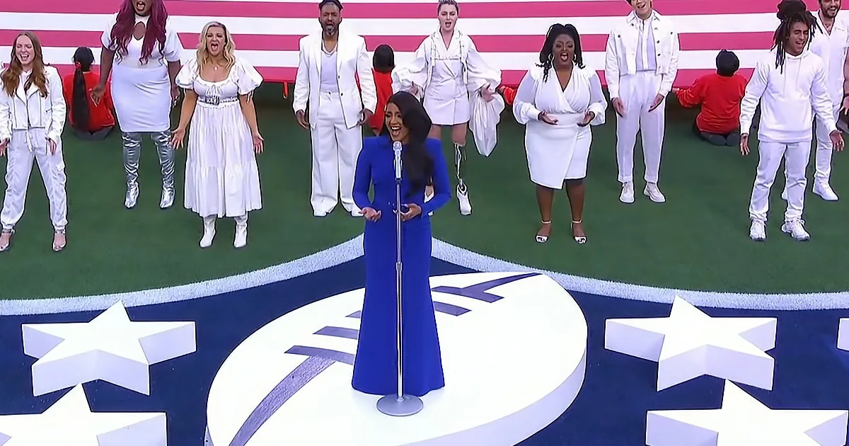 A woman in a blue dress sings into a microphone, surrounded by a group in white, with an American flag backdrop.