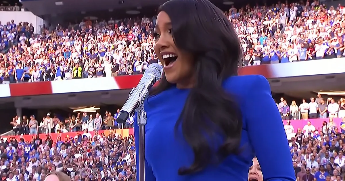 A woman in a blue dress sings into a microphone in front of a large crowd at a stadium.