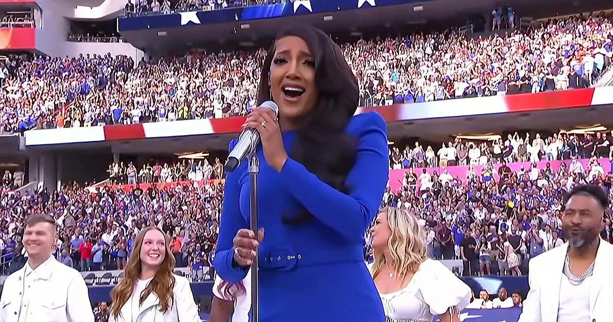 A woman in a blue dress sings into a microphone in front of a large, cheering crowd at a stadium.