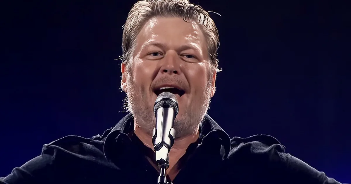 A man with a beard sings into a microphone, wearing a dark shirt, against a dark background.