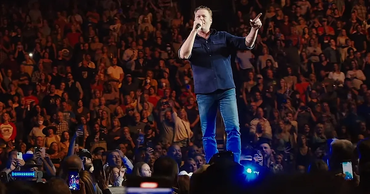 A man in a blue shirt and jeans stands on stage, singing into a microphone, with a large crowd behind him.