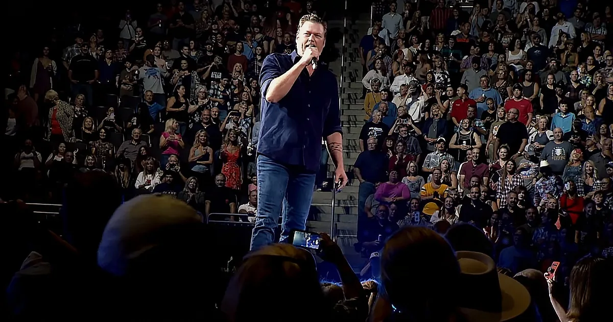 A man in a blue shirt sings into a microphone on stage, surrounded by a large, engaged audience.