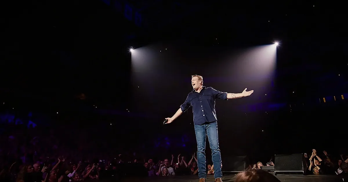 A man stands on stage with outstretched arms, illuminated by spotlights, surrounded by an enthusiastic crowd.