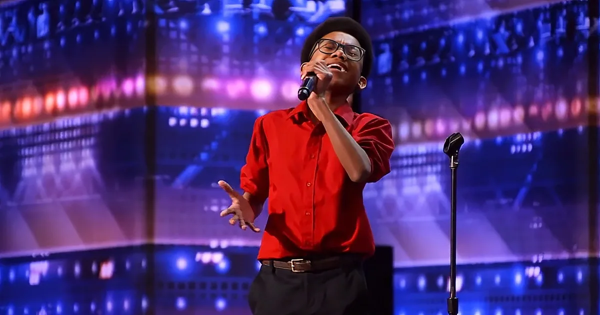 A person in a red shirt and black hat sings passionately on stage with a microphone.