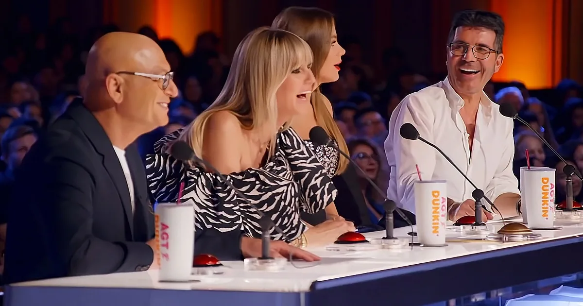Three judges at a table with microphones, smiling and engaged, in front of a blurred audience.