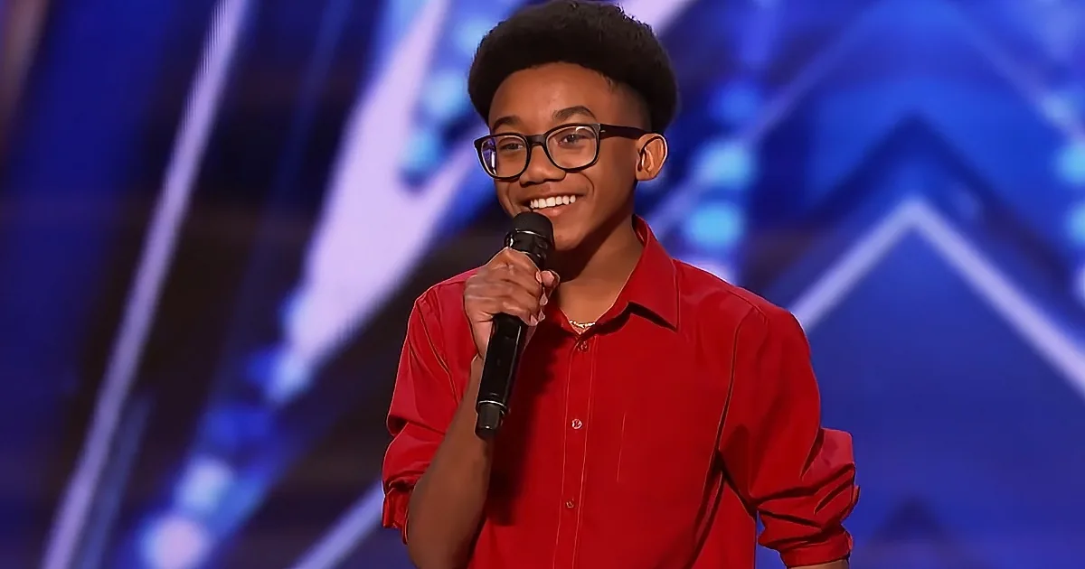 A young person in a red shirt holds a microphone, smiling on stage with a blue geometric background.