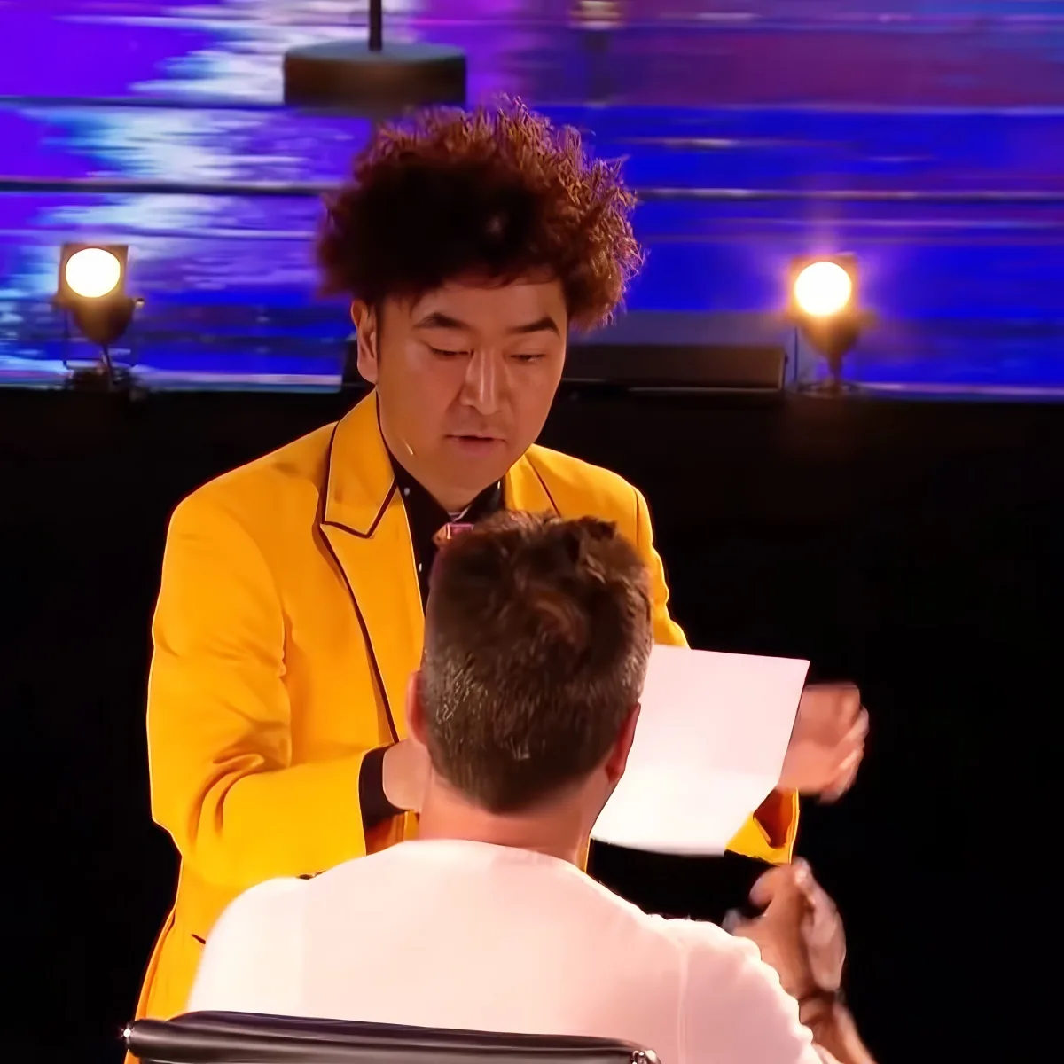 A man in a yellow suit hands a paper to another man in a white suit, with a stage backdrop and spotlights in the background.