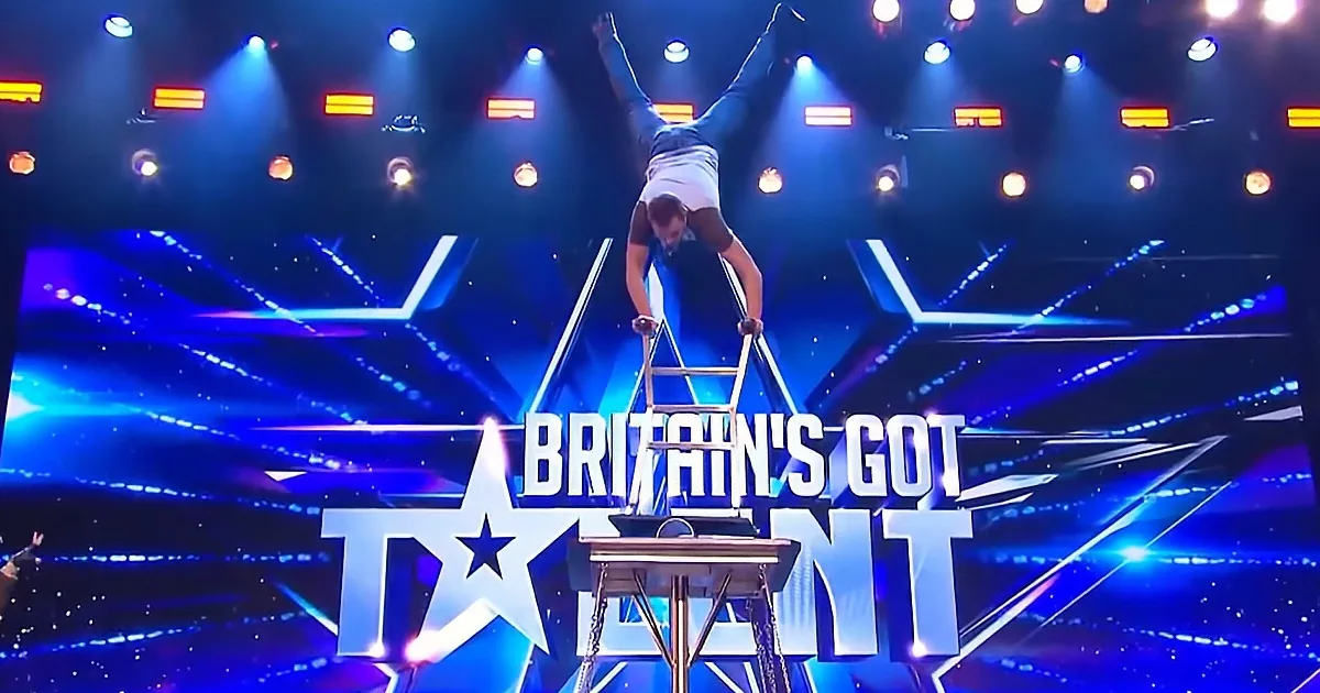 A performer balances on a ladder in a dramatic pose on a stage with "Britain's Got Talent" branding.