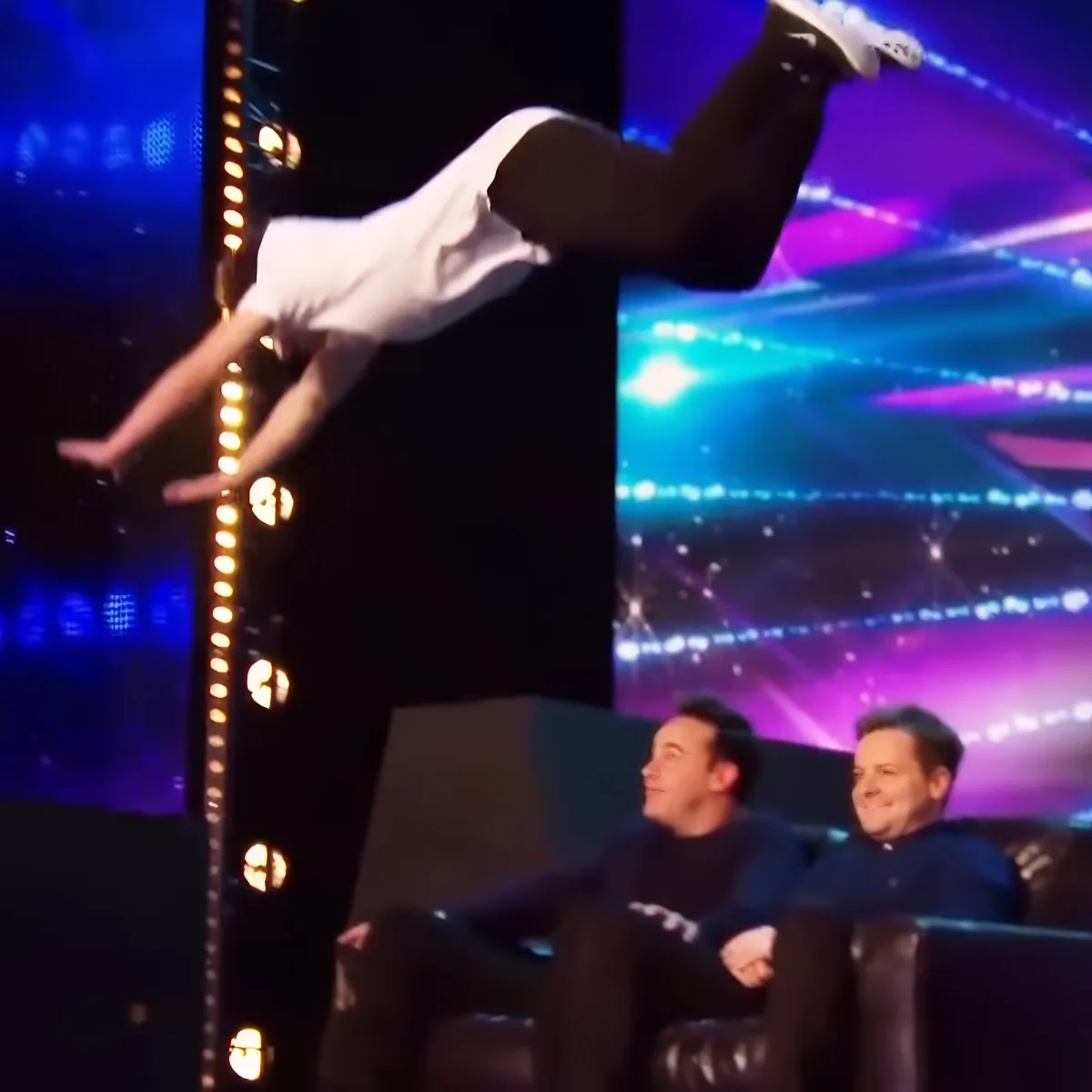 A person is performing an acrobatic move on a pole, with two men watching in the foreground.