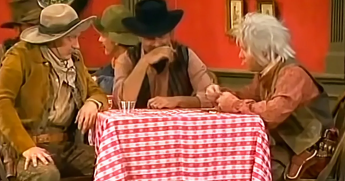 Four men in Western attire sit around a red-and-white checkered tablecloth, engaged in conversation.
