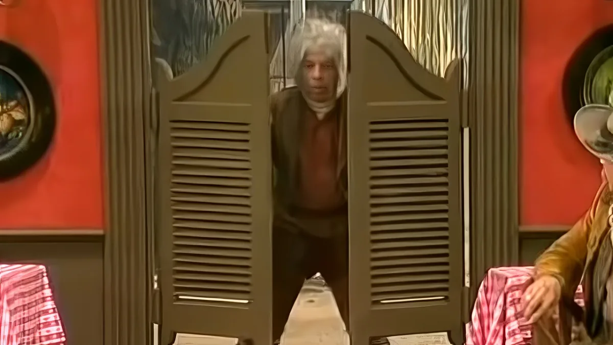 An older man with white hair stands behind a large wooden door in a room with red walls and decorative plates.