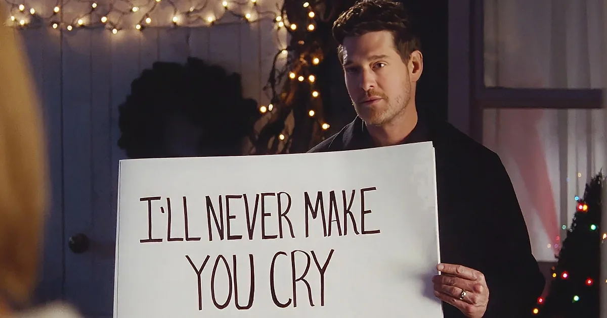 A man holds a sign saying "I'll never make you cry" in front of a decorated Christmas tree.