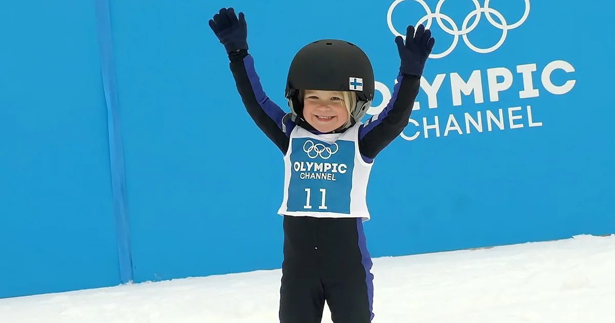 A child in ski gear celebrates with arms raised, wearing a helmet and jersey with "Olympic Channel" and number 11.