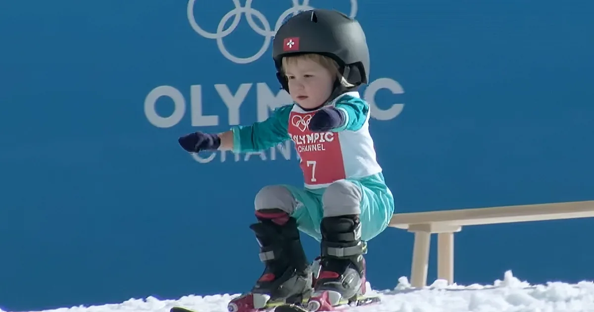 A child in ski gear sits on skis, pointing forward, with an Olympic backdrop.