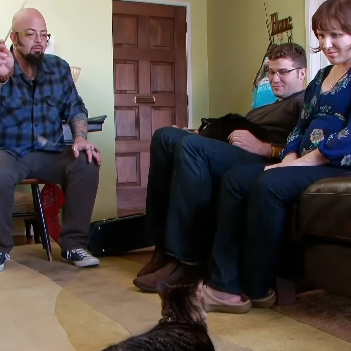 A man in a plaid shirt sits on a chair, gesturing, while two others sit on a couch, and a cat lies on the floor.
