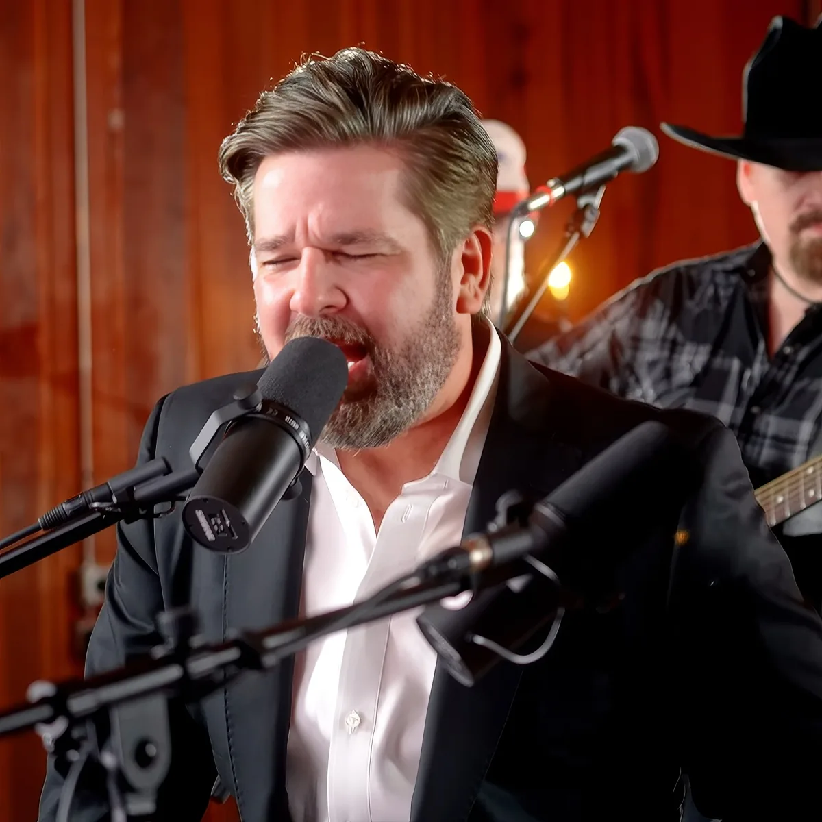 A man in a suit sings into a microphone, with another person playing guitar in the background.