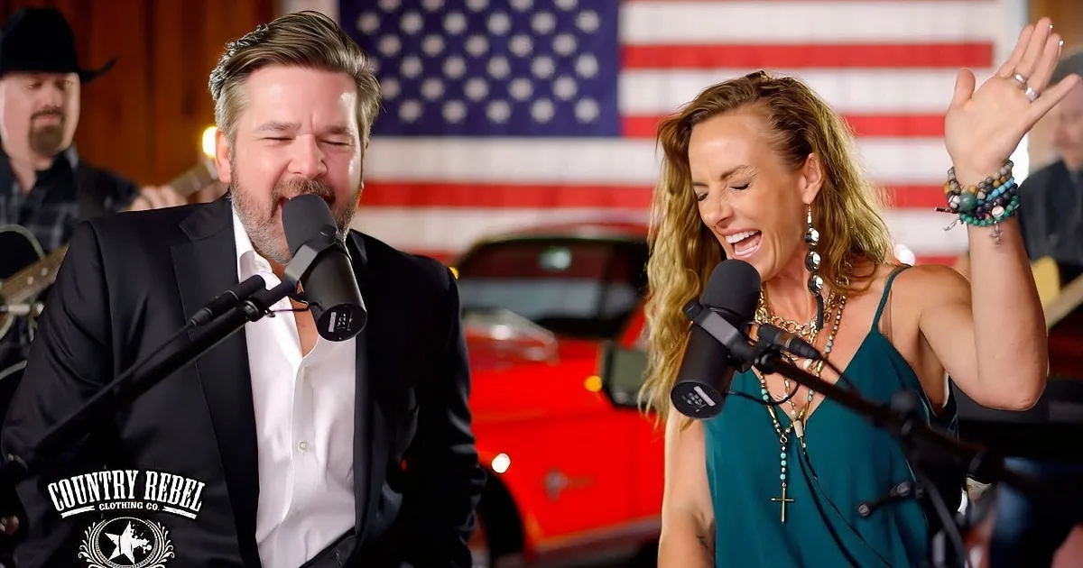 A man and woman sing into microphones, an American flag and musicians in the background.