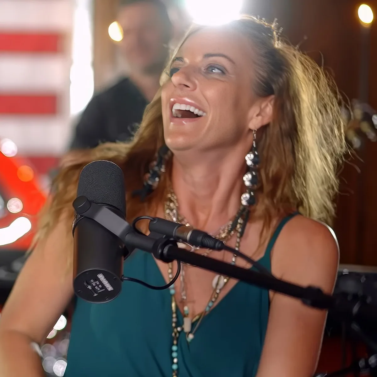 A woman with long blonde hair and earrings sings into a microphone, wearing a teal top and multiple necklaces.