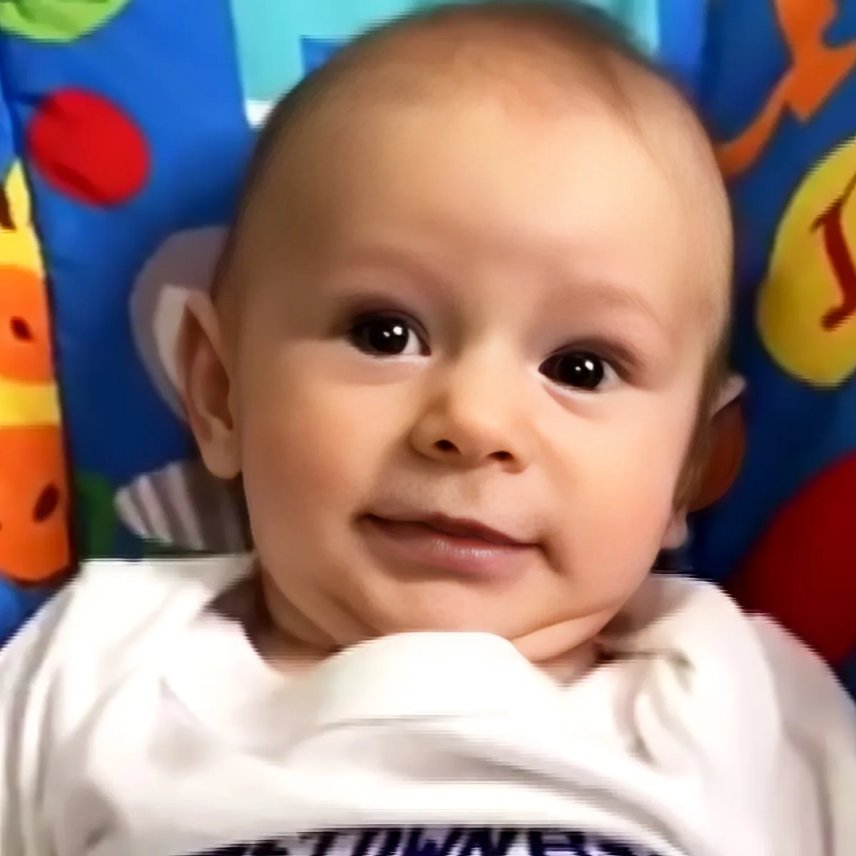 A baby with a bald head, wearing a white shirt, smiles in front of a colorful background with toys.