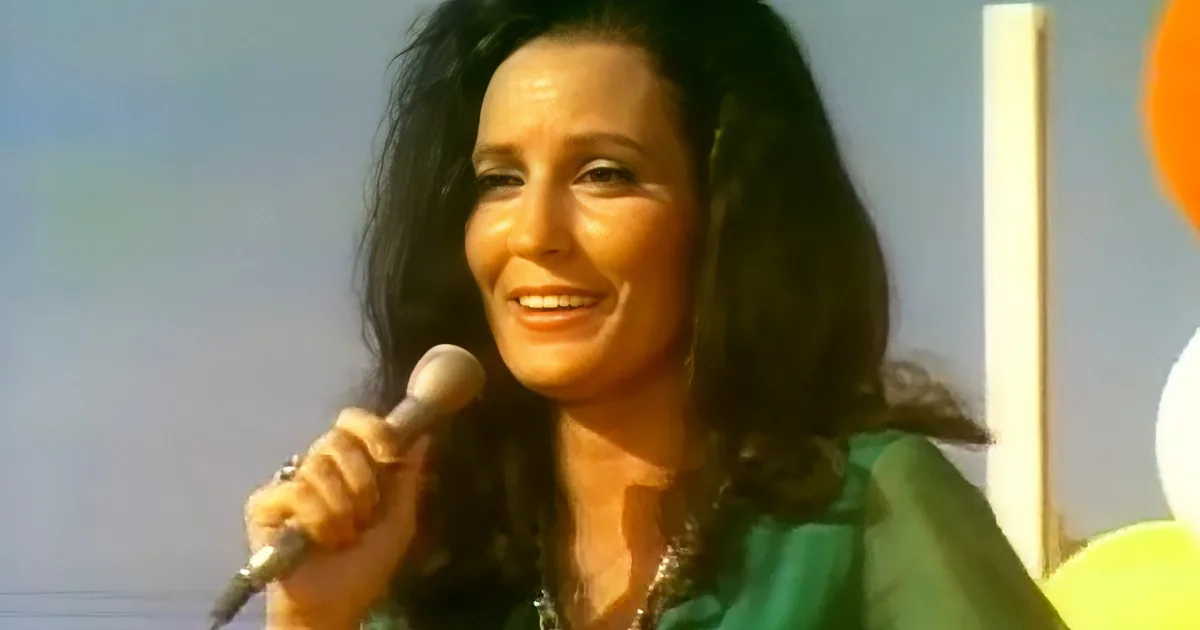 A woman with long dark hair smiles while holding a microphone, wearing a green top.