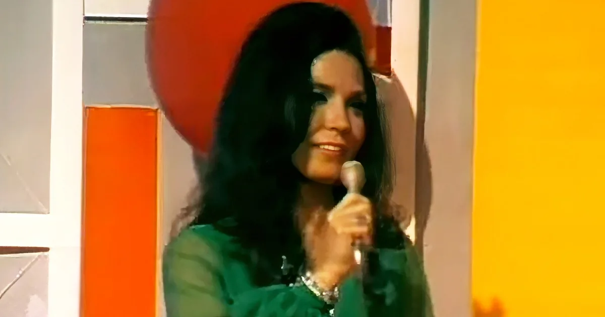 A woman with long dark hair in a green outfit holds a microphone, standing in front of a colorful background.