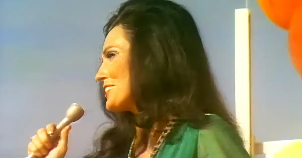 A woman with long dark hair holds a microphone, wearing a green top, against a blurred background.