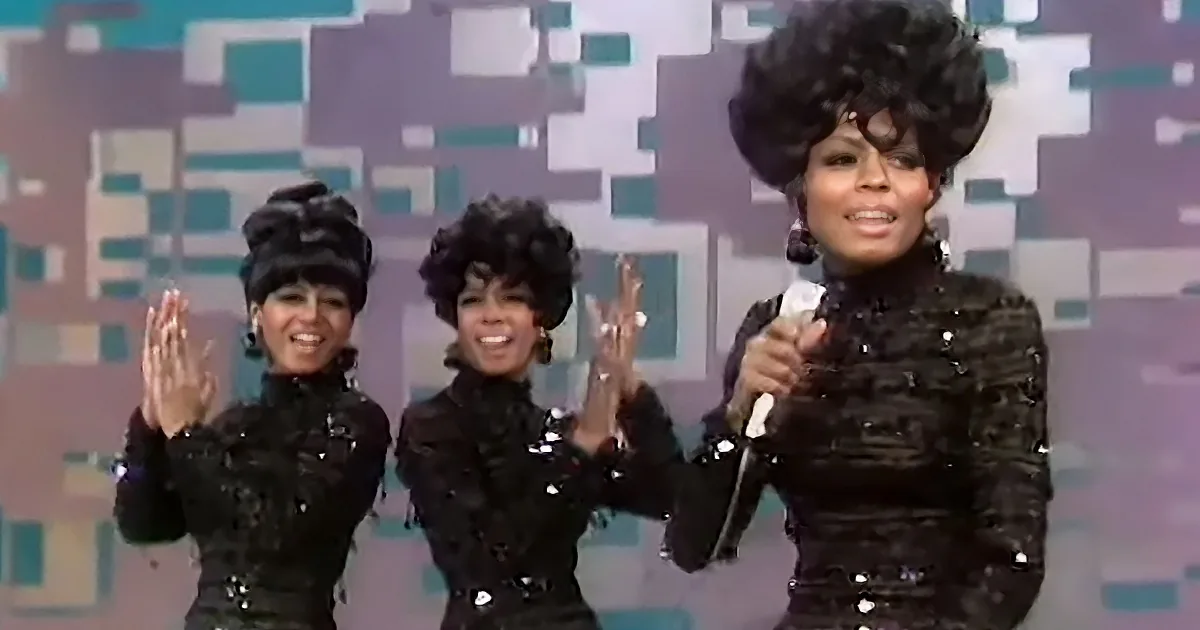 A woman in a black sequined outfit holds a microphone, smiling and clapping with two others in matching outfits.