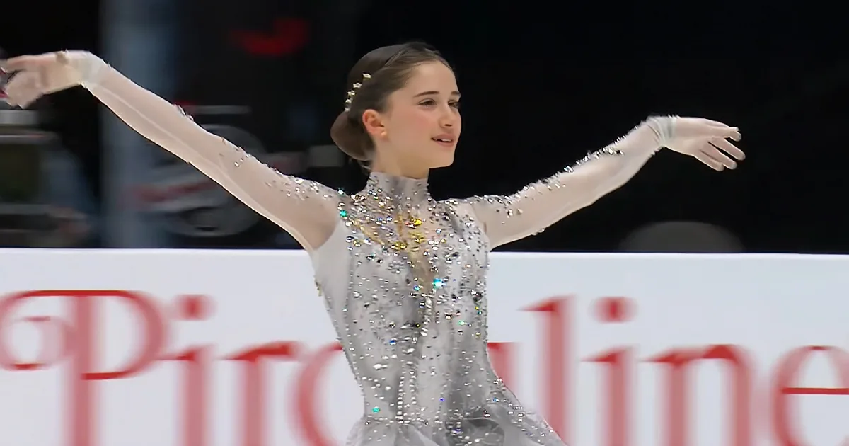 A figure skater in a sparkling silver dress with arms outstretched on the ice.