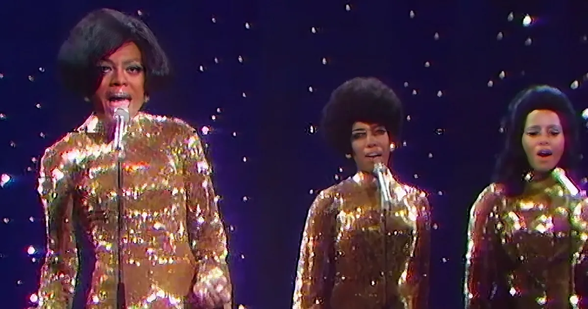 A performer in a glittery gold outfit sings into a microphone, accompanied by two others in similar attire.