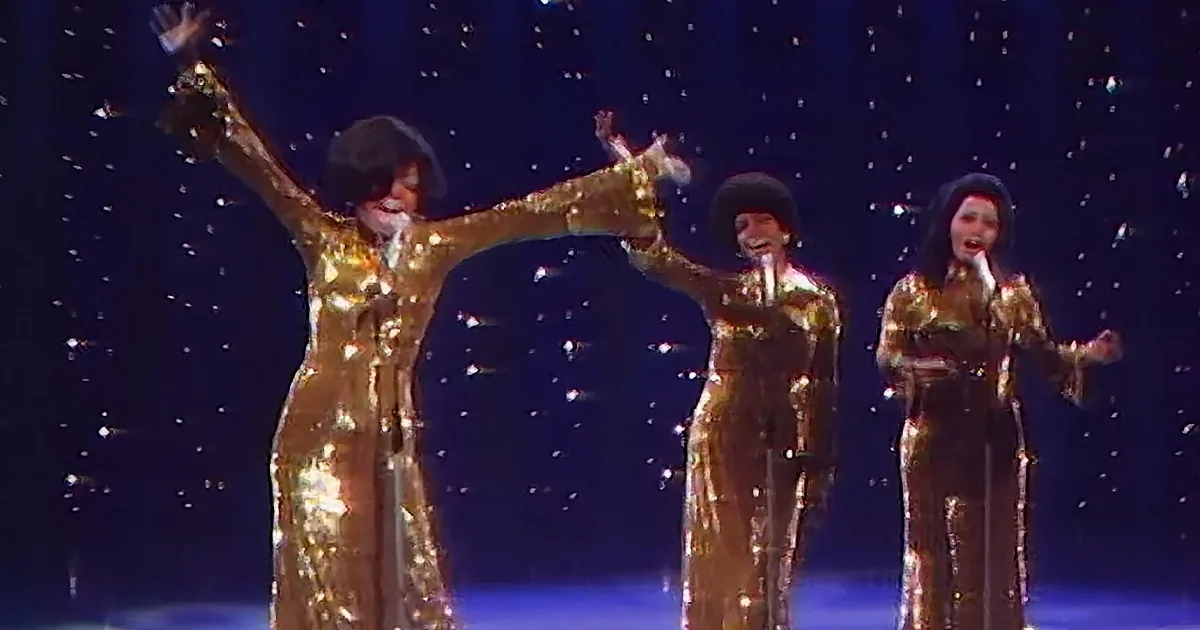 A performer in a shiny gold outfit raises their arms, surrounded by other performers in similar attire, against a starry...
