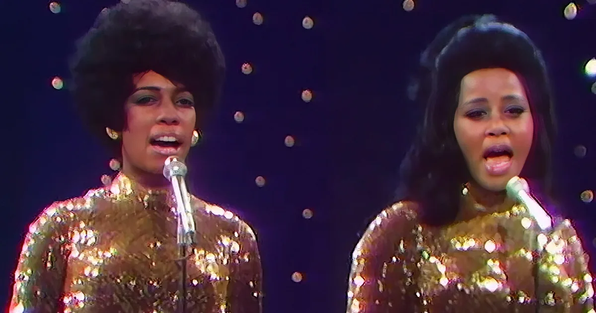 Two women in shimmering gold outfits sing into microphones against a starry backdrop.
