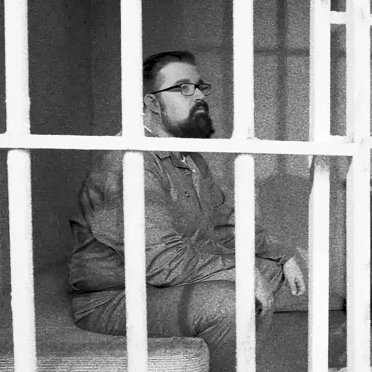 A man with glasses and a beard sits behind bars, wearing a button-up shirt and pants.