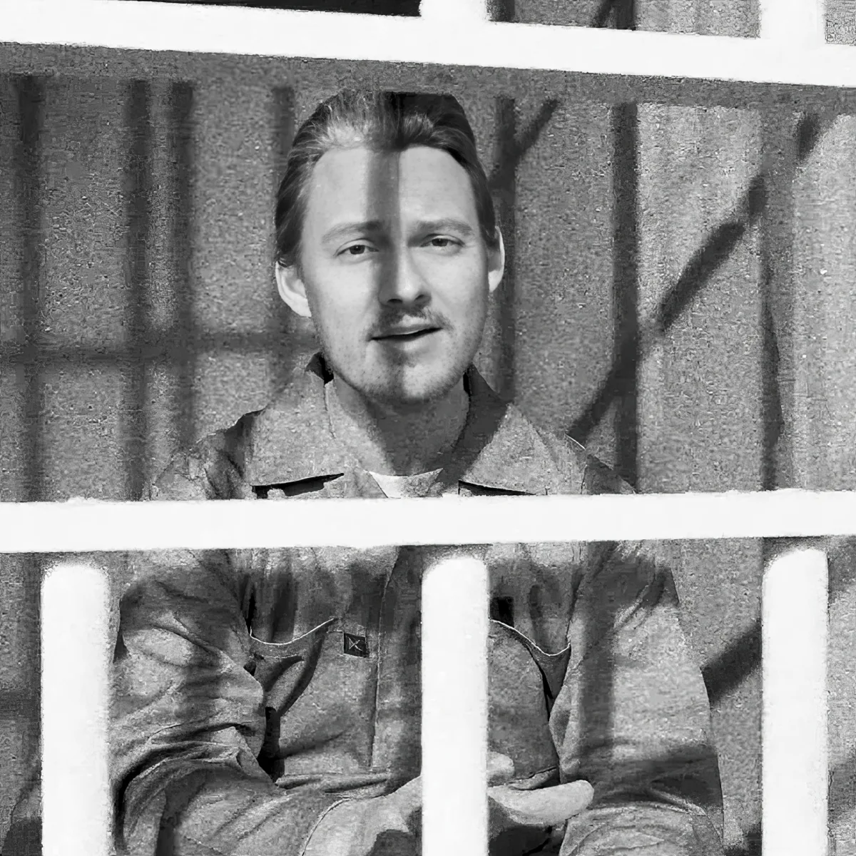A man in a prison uniform sits behind bars, looking directly at the camera.