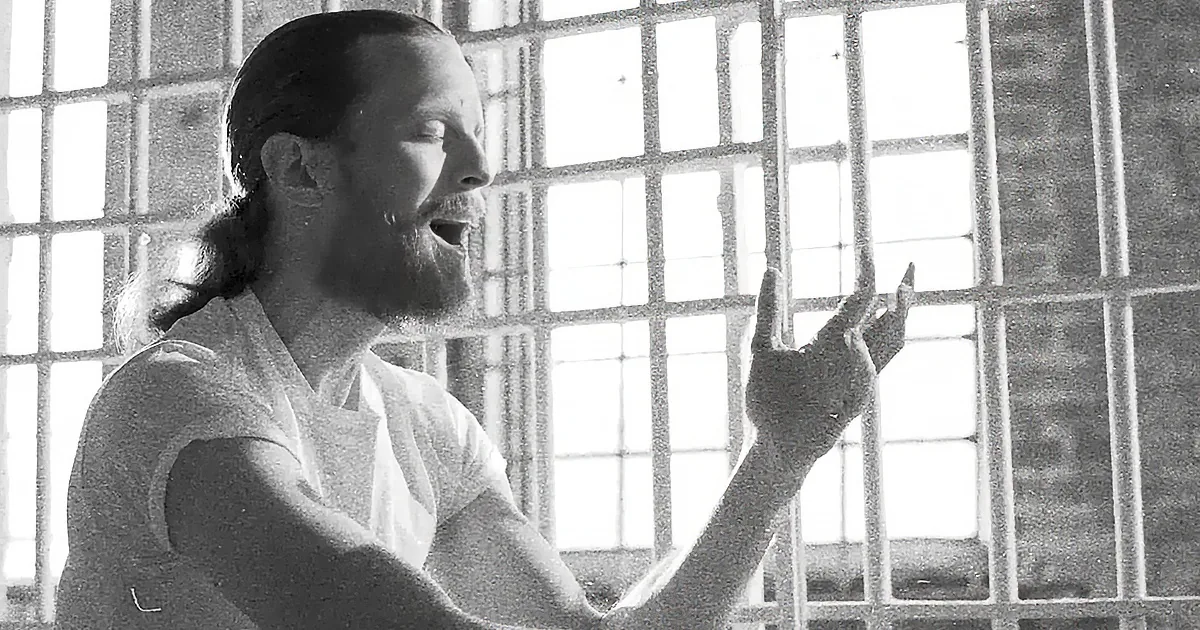 A man in a white shirt stands in front of a barred window, gesturing with his right hand.