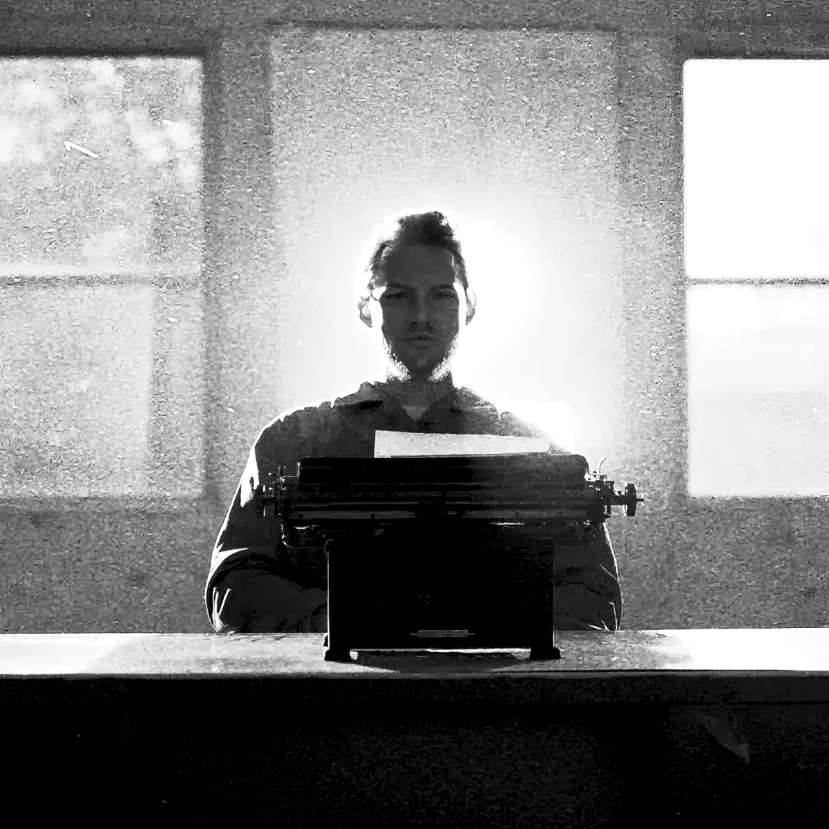 A person sits at a desk with a typewriter, illuminated by light from a window behind them.
