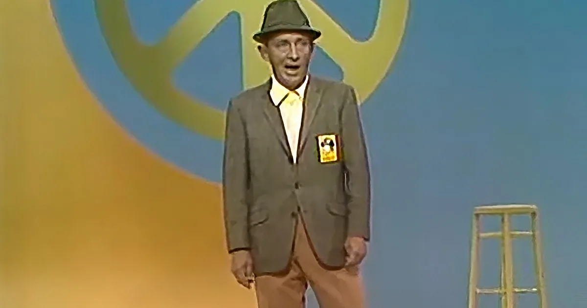 A man in a suit and hat stands on stage with a peace sign backdrop.