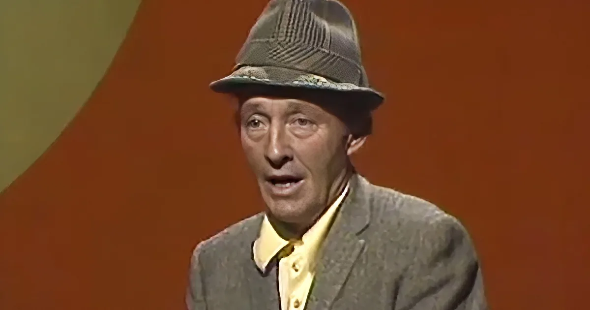An older man wearing a patterned hat and a gray suit jacket with a yellow shirt underneath is speaking.