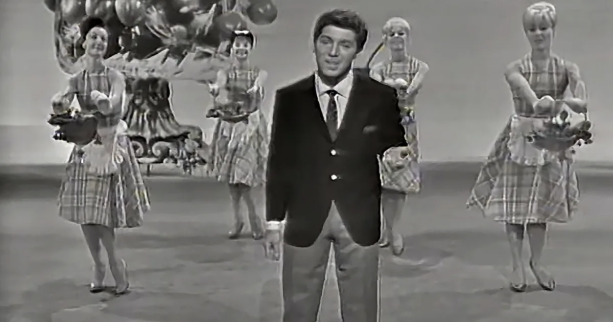 A man in a suit stands center stage, surrounded by women in plaid dresses holding baskets.