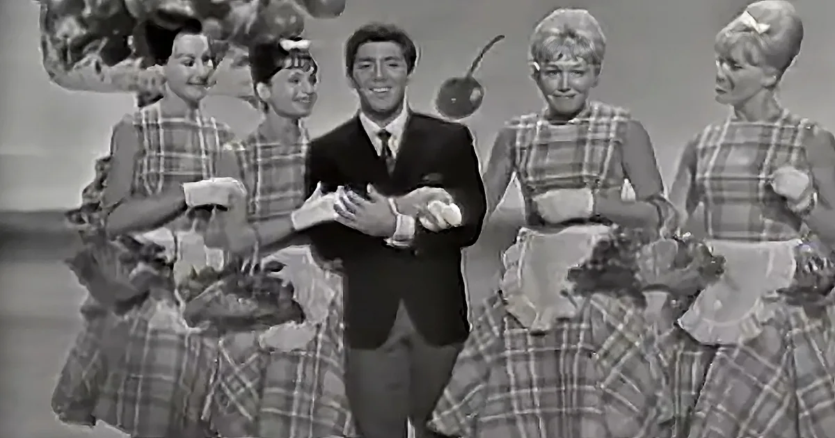 A man in a suit stands center stage, surrounded by women in plaid dresses, holding flowers.