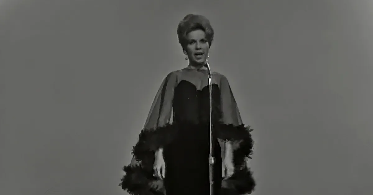 A woman in a black dress with a feathered cape stands in front of a microphone, singing.